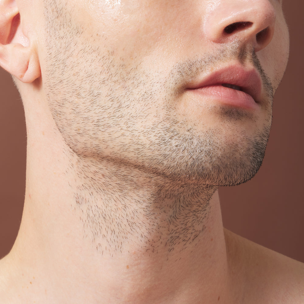 Close-up of a man's lower face, neck, and upper chest showing natural stubble and facial hair against a warm brown background.